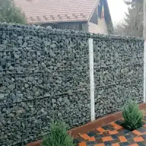 A sturdy, gray stone-filled gabion fence with white posts lines a checkered patio. Small green shrubs add contrast, while a house roof is visible behind.