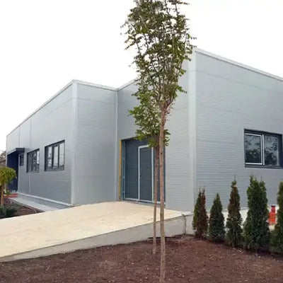 Modern single-story gray modular building with large windows, surrounded by small trees and shrubs, under a clear blue sky, conveys simplicity and functionality.