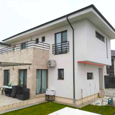Modern two-story house with white and beige exterior, flat roof, and balcony. The patio features dark furniture and a small sink, creating a cozy atmosphere.